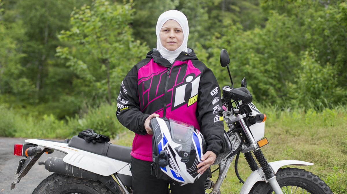 A woman, wearing a white hijab and motorcycle jacket, stands in front of her motorcycle holding a helmet.